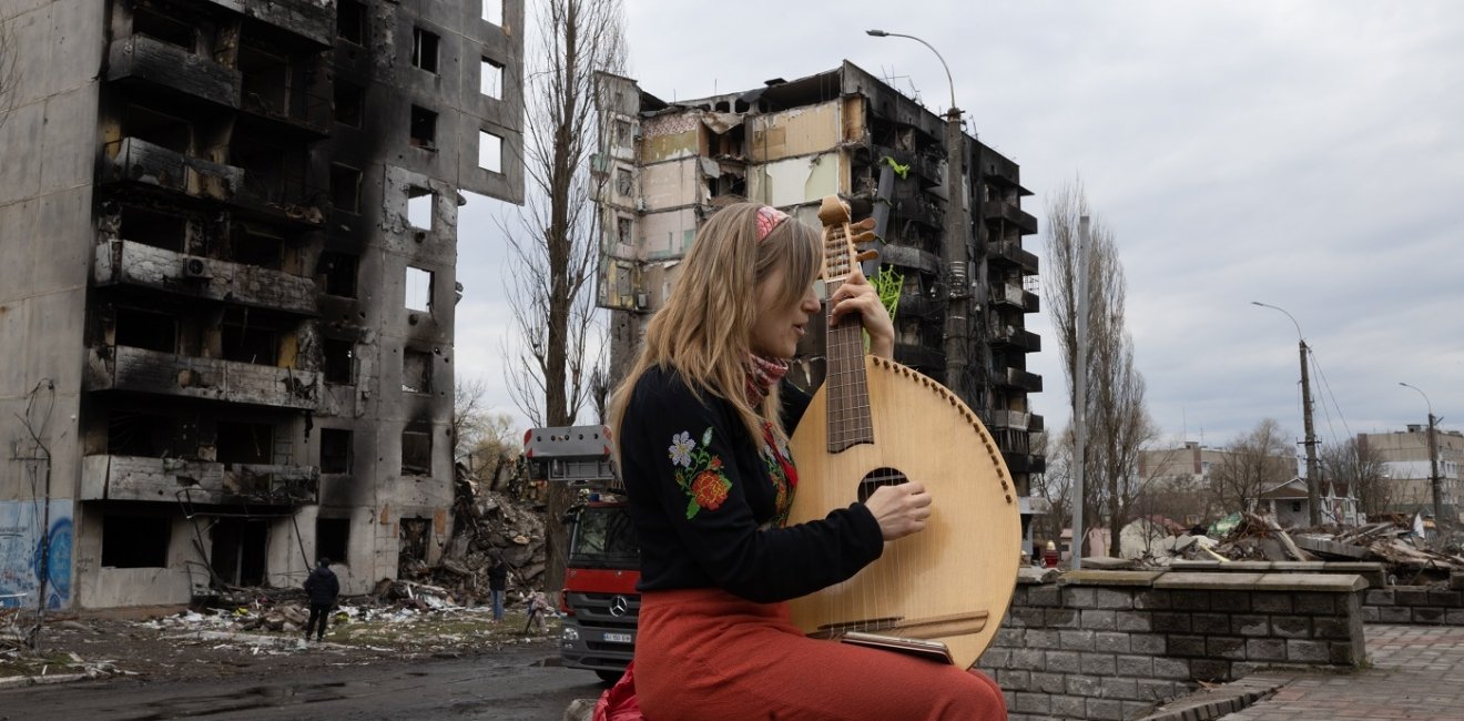 A woman plays a stringed instrument in front of a destroyed building in Bodoryanca, Kyiv Oblast, Ukraine