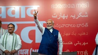 Narendra Modi stands in front of a red background, holding up his hand in a victory sign.