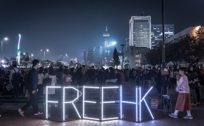 A group of protestors are gathered at night, lights spell out Free HK in front of them.