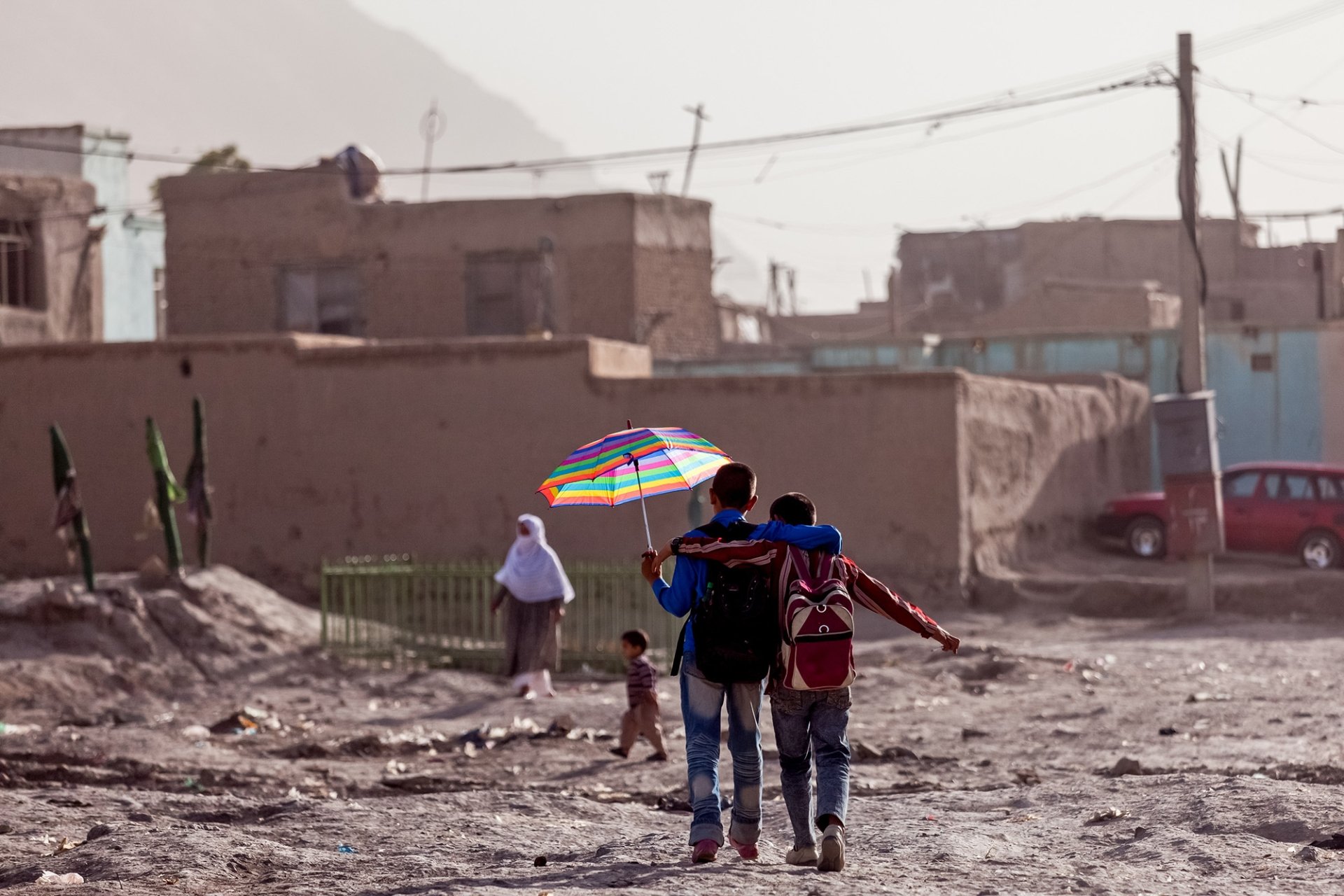 Two young people walk arm in arm along a dirt path, one of them holding an umbrella above both of their heads.